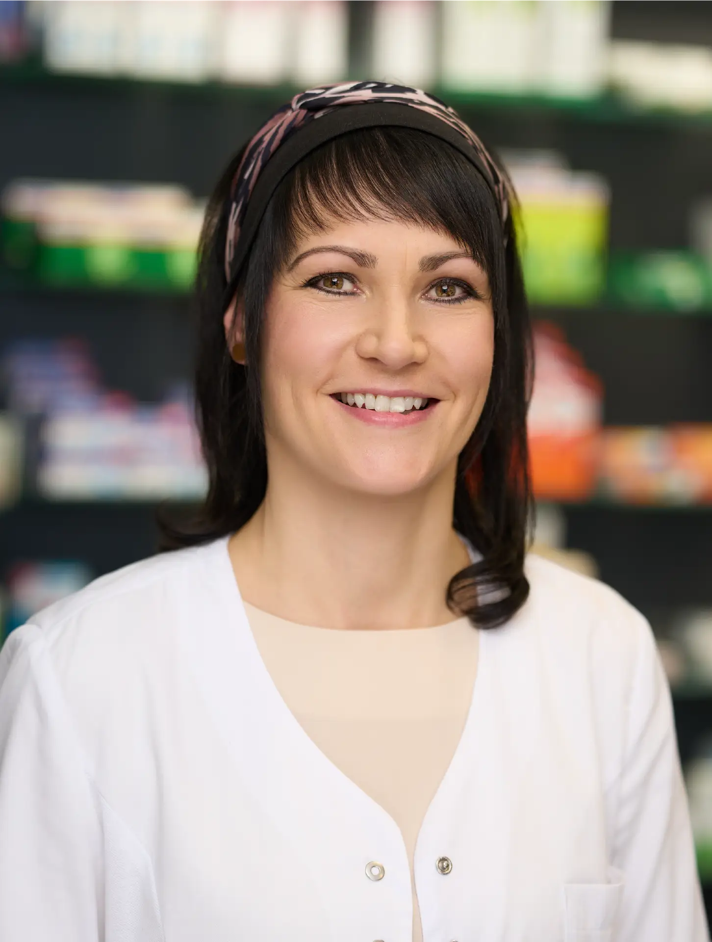 Freundliches Teamportrait einer Apothekerin aus der Rhön mit dunklen Haaren und Stirnband, im weißen Kittel, vor unscharfem Hintergrund.